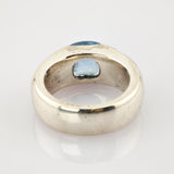 Silberner Ring mit breitem Band und großem facettiertem blauem Edelstein auf reflektierender weißer Oberfläche.