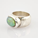 Silberring mit großem ovalem grün-blau schimmerndem Edelstein auf reflektierender weißer Oberfläche