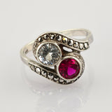 Eleganter Silberring mit zwei großen Steinen, klar und tiefrot, geschwungenes Metallband mit kleinen schwarzen Steinen.