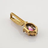 Kleiner Anhänger aus goldfarbenem Metall mit facettiertem lila und pinkem Edelstein auf weißem, spiegelndem Untergrund.
