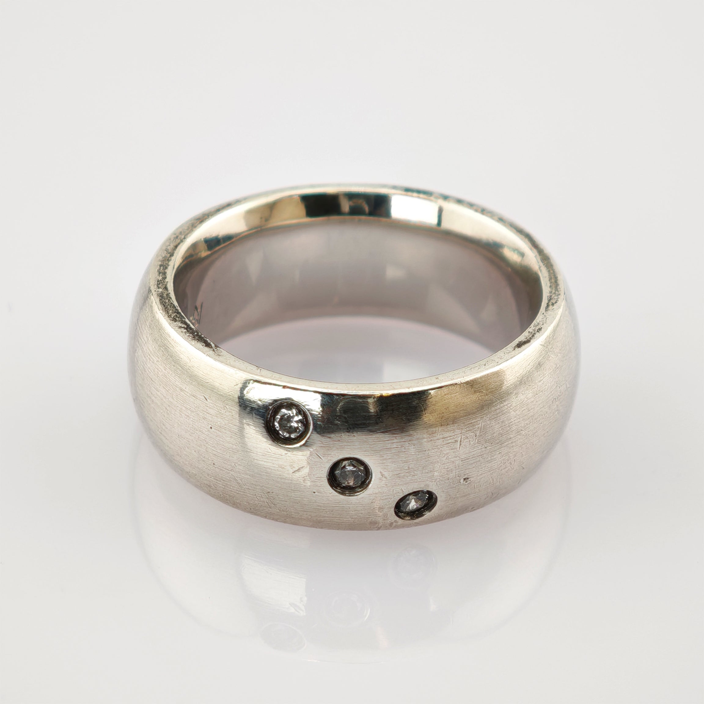 Breiter Silberring mit drei kleinen klaren diagonal angeordneten Edelsteinen auf glänzender Oberfläche.