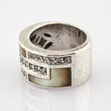 Breiter Silberring mit geometrischen Einlagen aus Perlmutt und kleinen klaren Steinen auf reflektierender Oberfläche