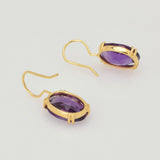 Elegante Goldohrringe mit ovalen, facettierten violetten Edelsteinen in Goldfassung auf weißem Hintergrund.