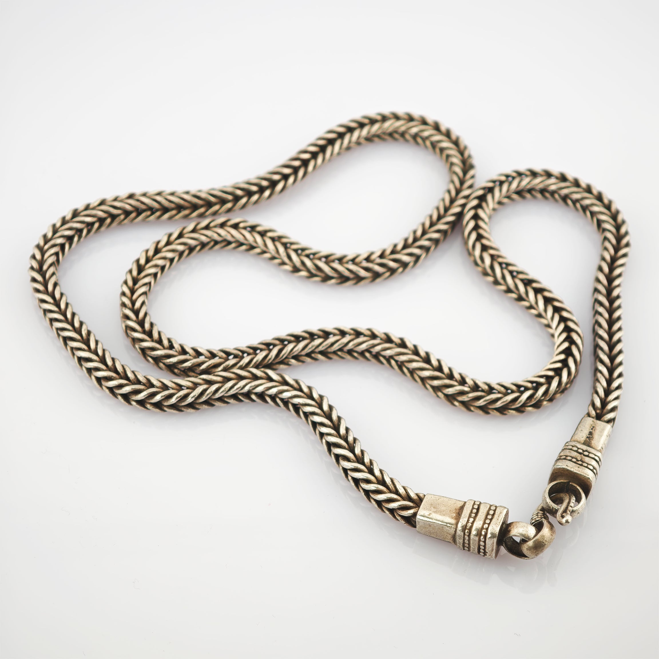 Geflochtene metallische Halskette mit strukturierten Endstücken und Hakenverschluss vor weißem Hintergrund