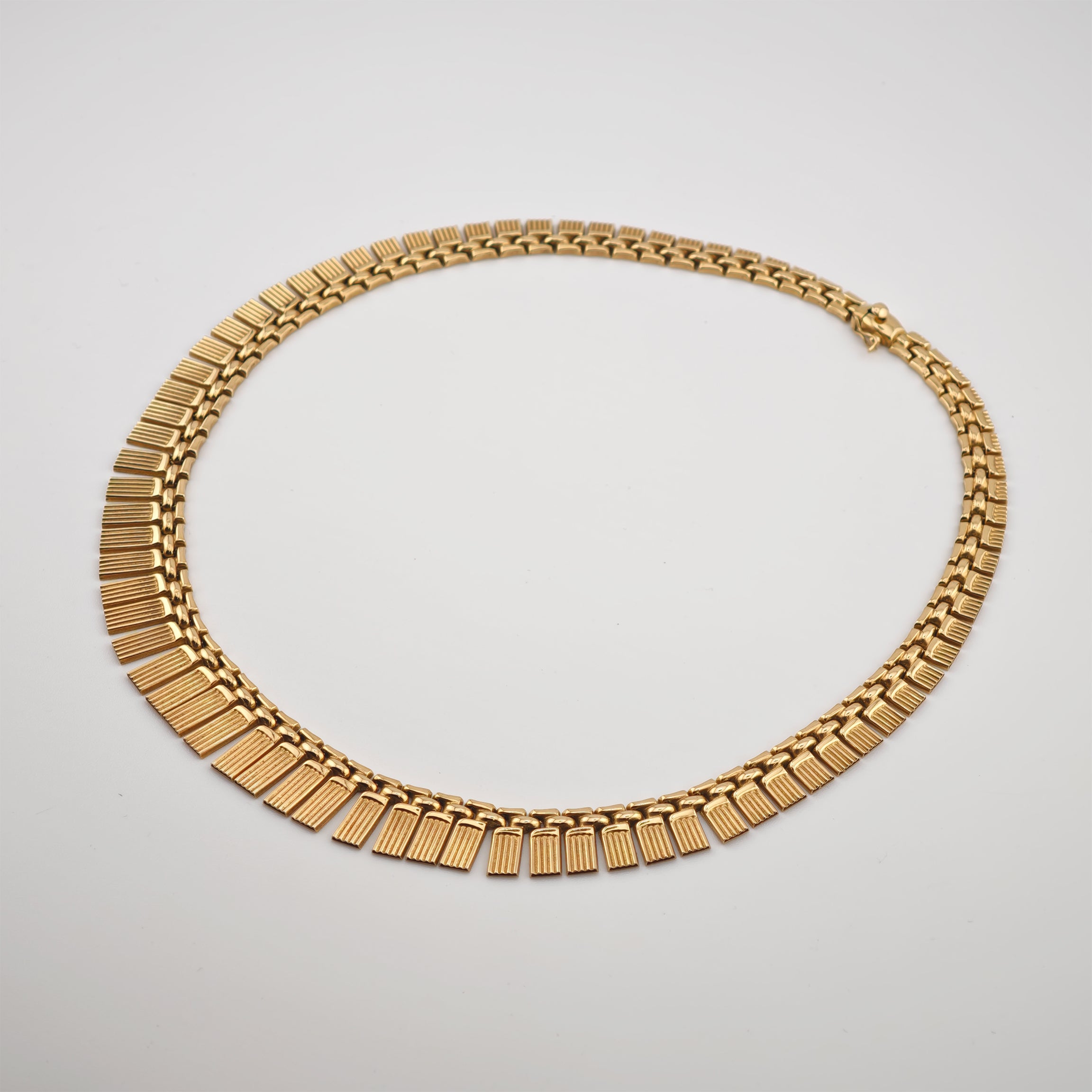 Goldene Halskette mit rechteckigen, gerillten Gliedern und feinen Kettensegmenten auf hellem Hintergrund.