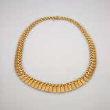 Goldene Halskette mit rechteckigen und quadratischen Gliedern auf hellgrauem Hintergrund.