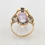 Goldener Ring mit ovalem lila Edelstein und filigraner Goldfassung auf reflektierendem Untergrund