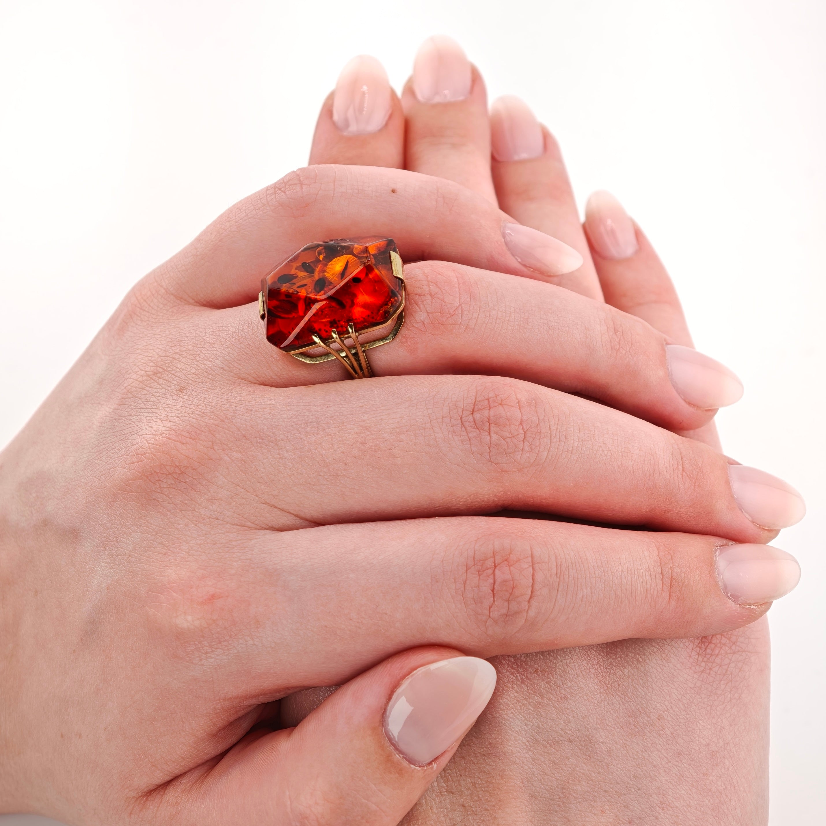Hand mit großem facettierten bernsteinfarbenen Ring auf Fuß mit gepflegten, lackierten Nägeln vor weißem Hintergrund.
