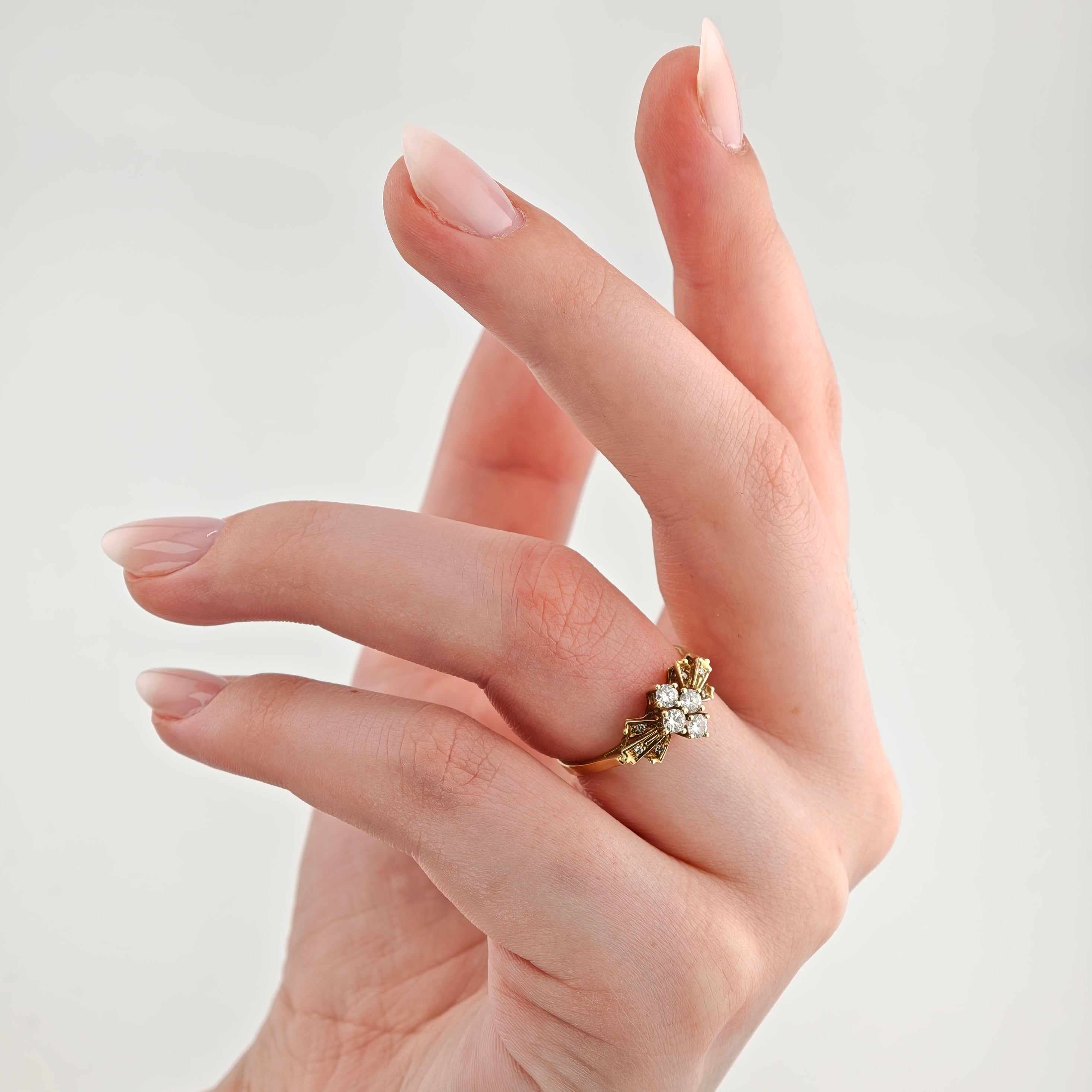 Hand mit ovalen, natürlich lackierten Nägeln und goldenem Ring mit Diamantcluster vor grauem Hintergrund