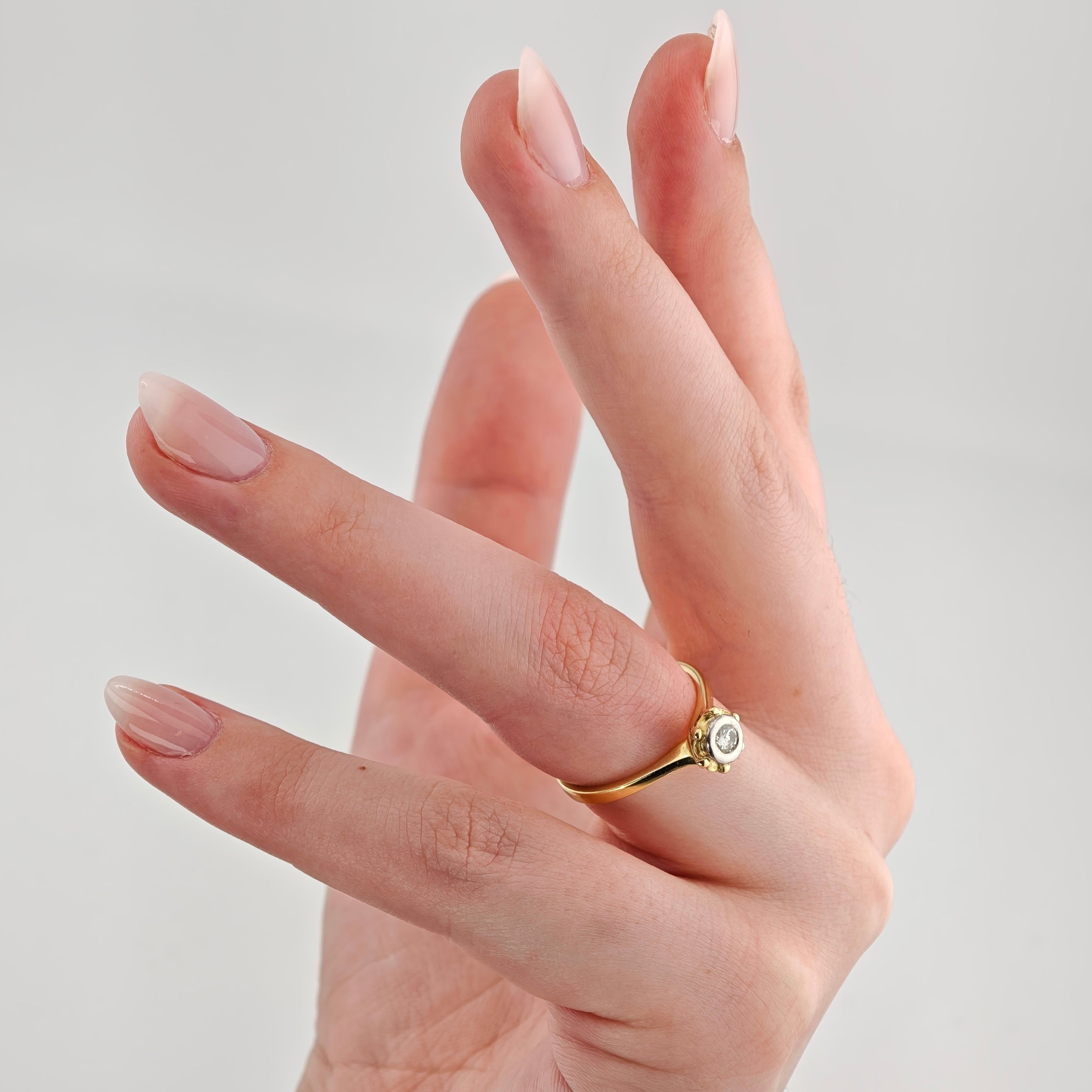 Linke Hand mit zartem goldenen Blumendamenring und klarem Edelstein vor grauem Hintergrund