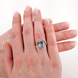 Nahaufnahme einer linken Hand, die einen goldenen Ring mit einem großen kissenförmigen hellblauen Edelstein und kleinen Diamanten trägt.