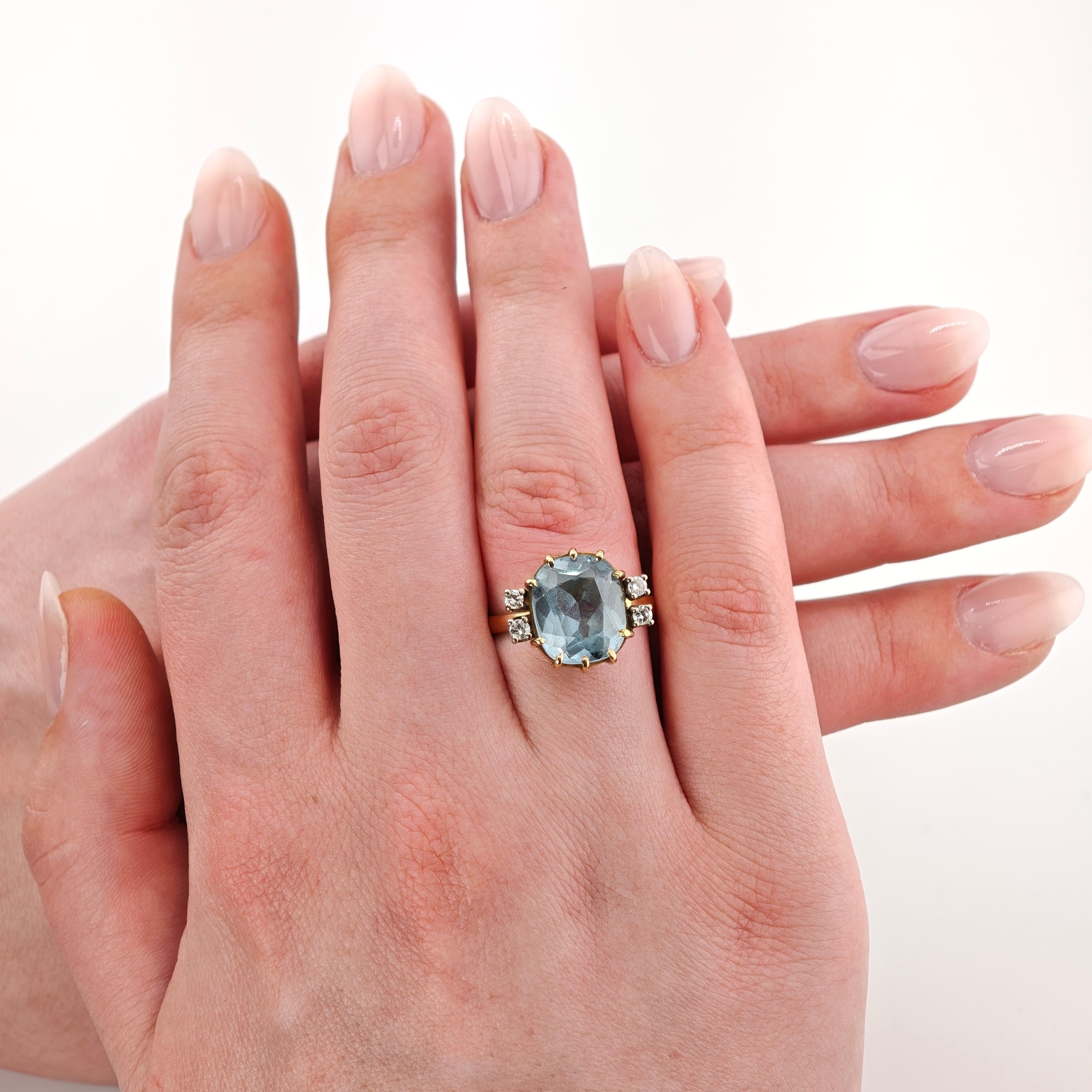 Nahaufnahme einer linken Hand, die einen goldenen Ring mit einem großen kissenförmigen hellblauen Edelstein und kleinen Diamanten trägt.