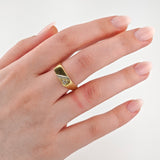 Linke Hand mit hellrosa lackierten Nägeln trägt einen goldenen Ring mit rechteckiger Platte und Diamant.