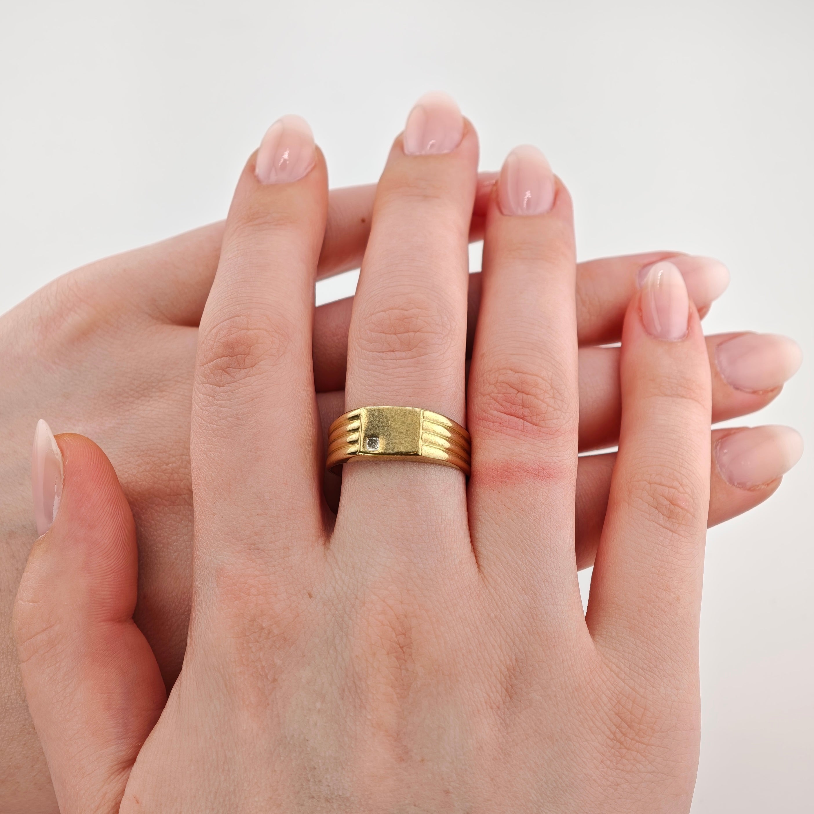 Linke Hand mit Goldring und kleinem Diamant am Ringfinger vor neutralem hellgrauem Hintergrund.