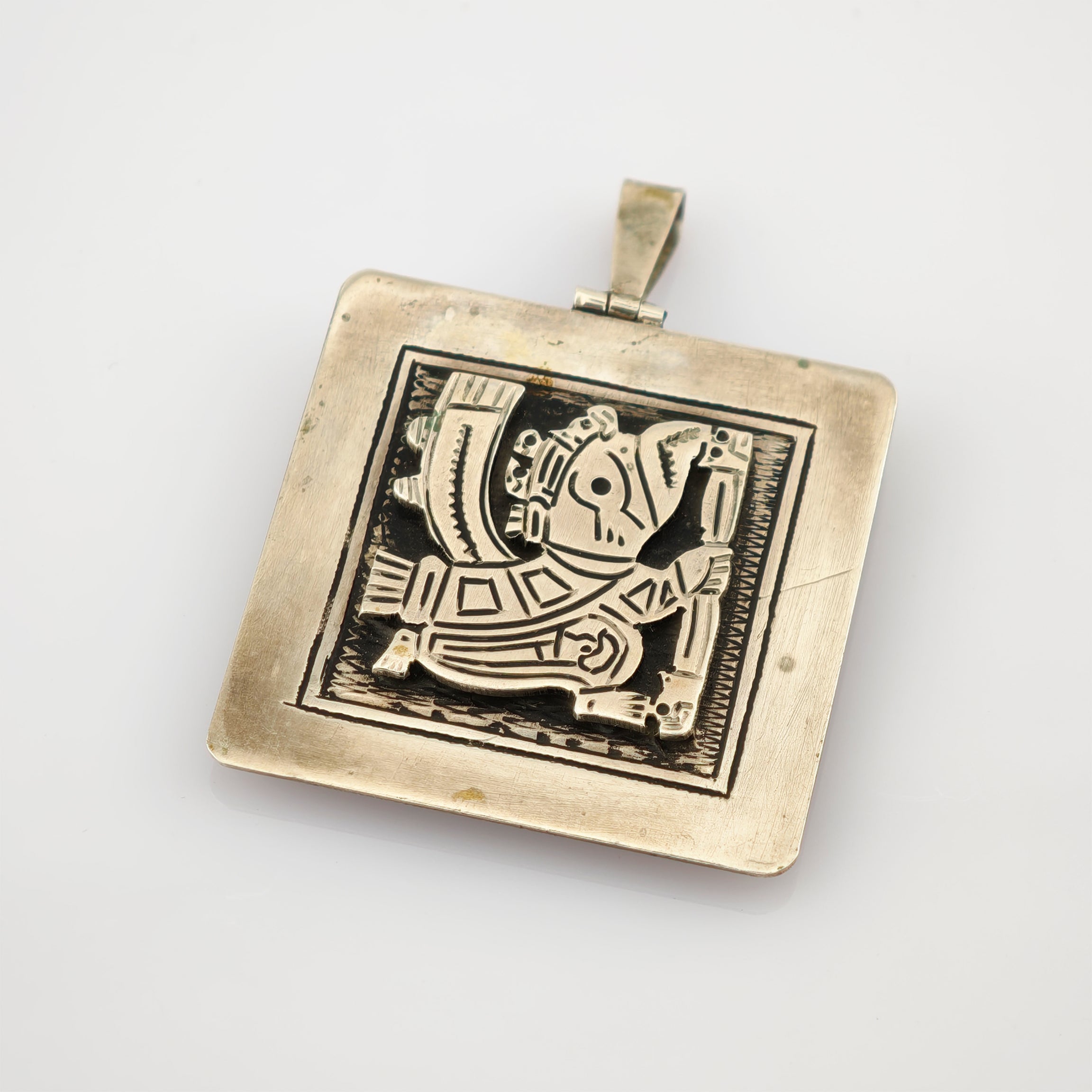 Quadratischer Metallanhänger mit leicht angelaufener Oberfläche und stilisierter Figur im Rahmen