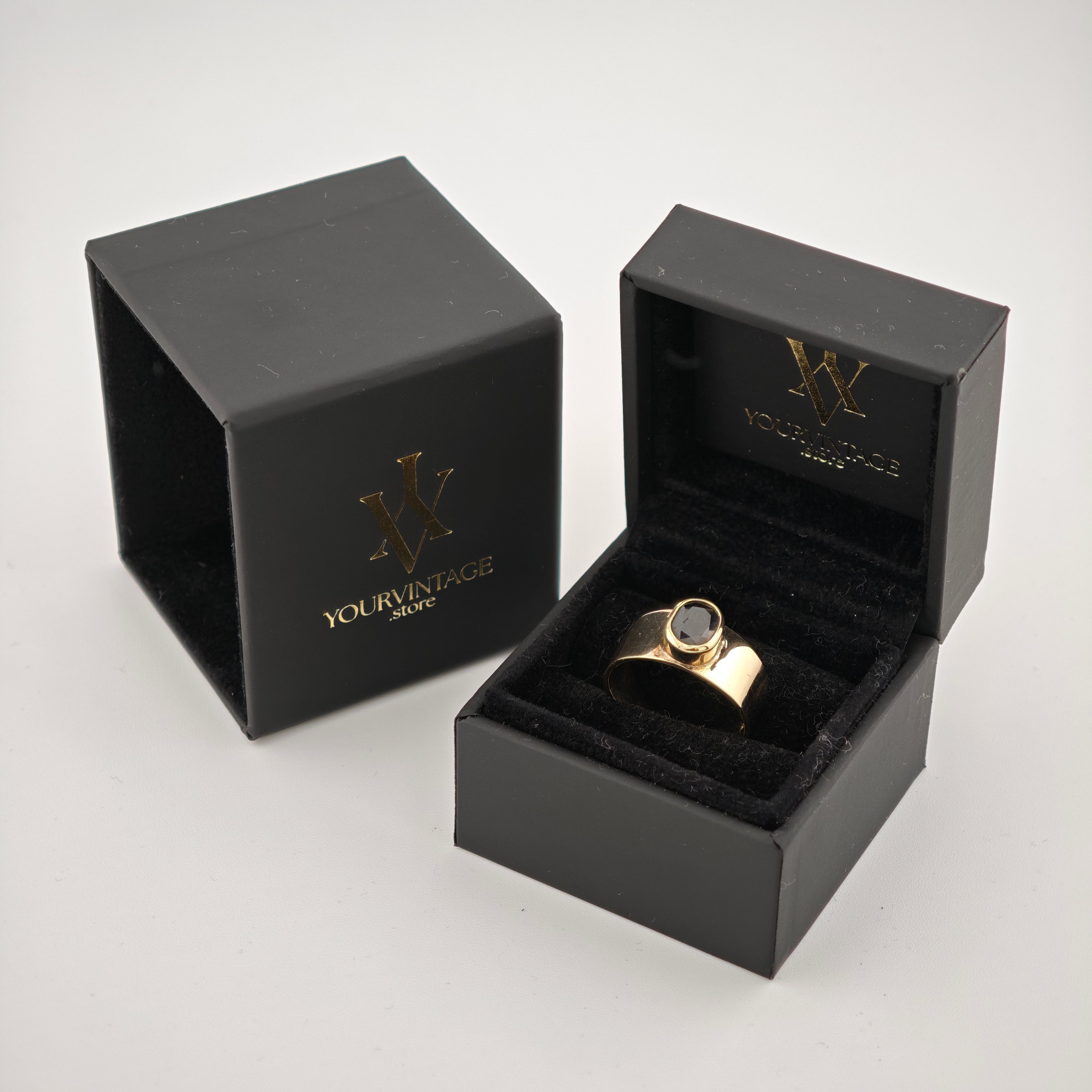 Zwei schwarze Schmuckschatullen mit goldenem Logo, eine offen mit breitem Goldring und dunklem Edelstein.