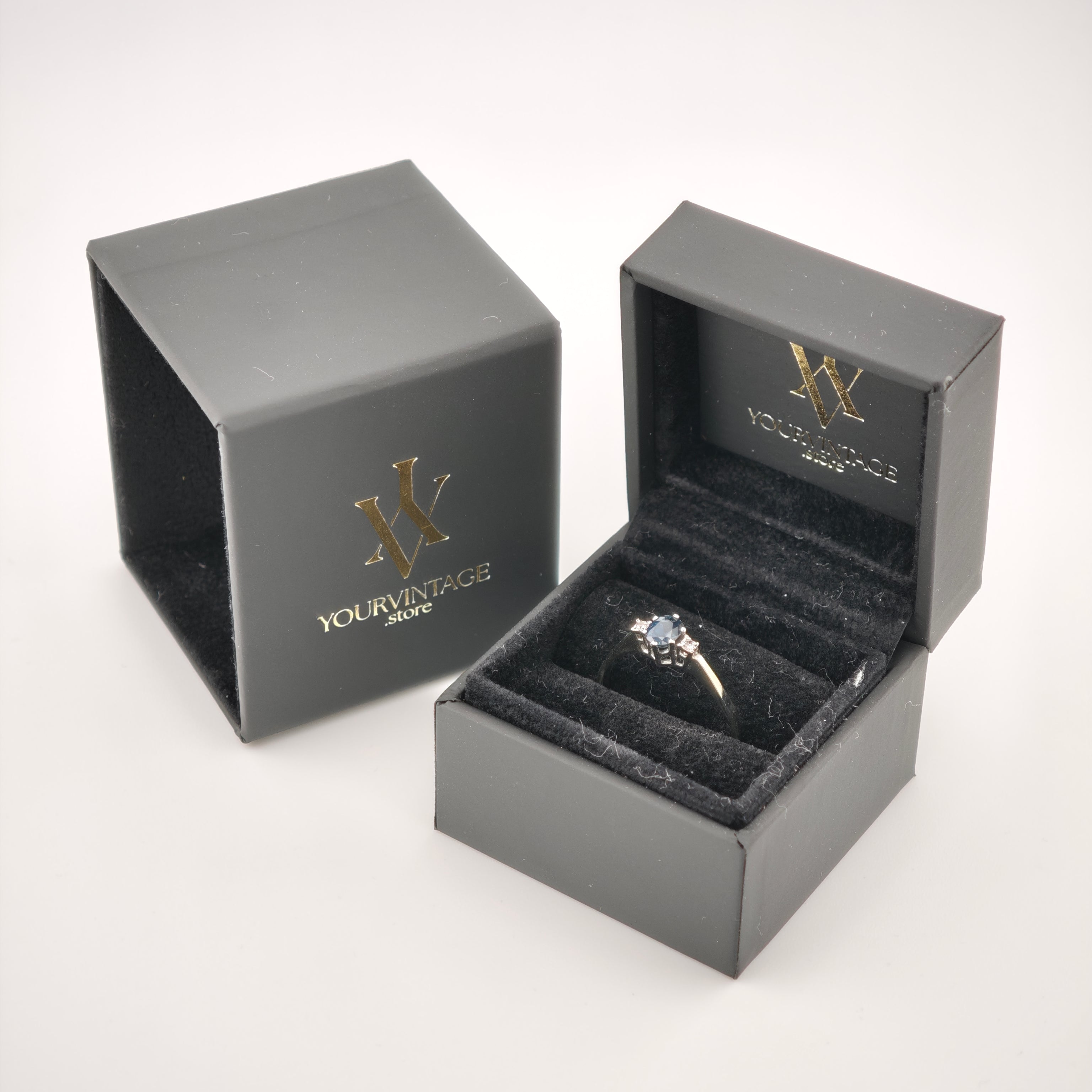 Schwarze Ringbox mit Samtfutter zeigt goldenen Ring mit dunklem Stein und zwei kleinen Seitsteinen, IhrVintage Store Logo.