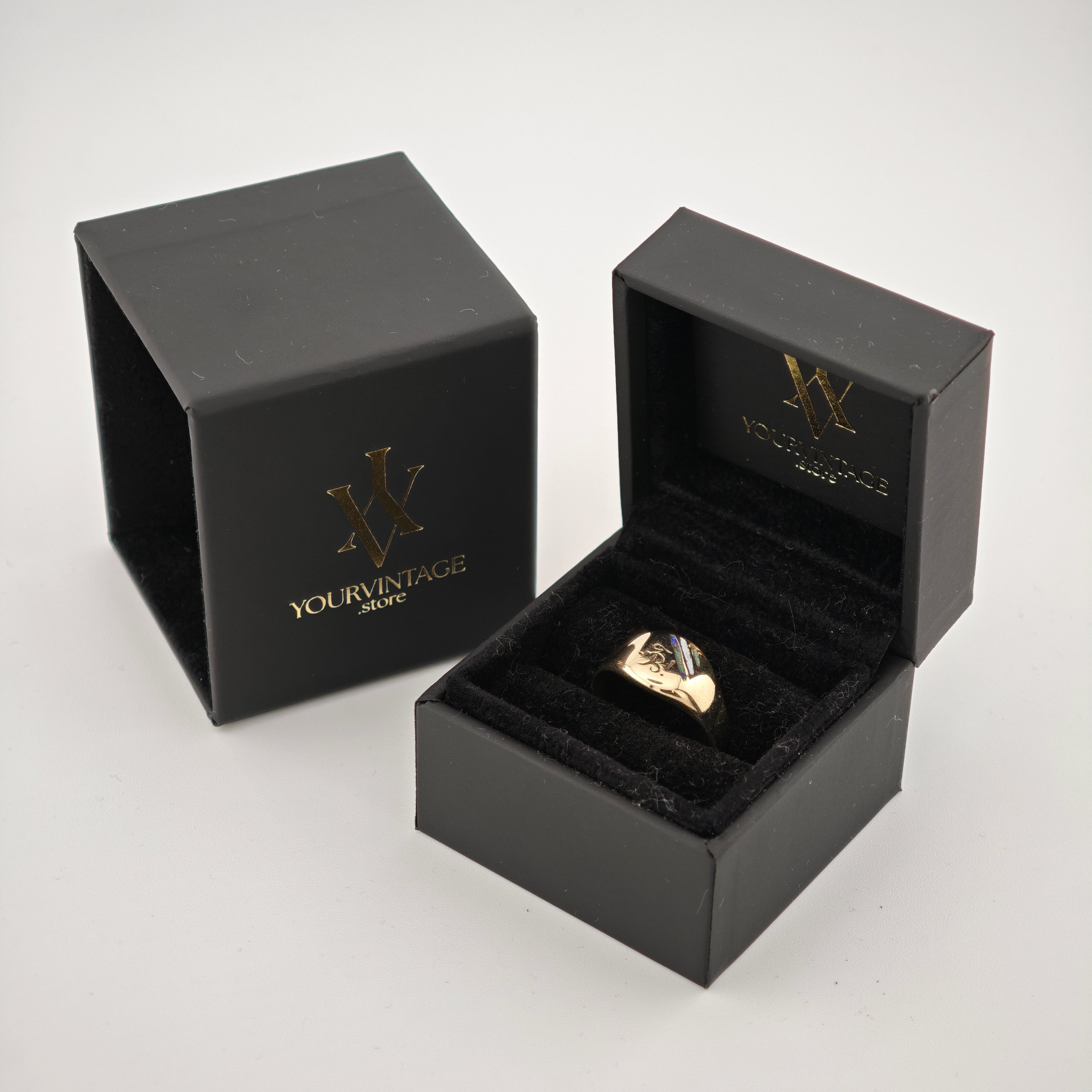 Offene schwarze Samt-Schmuckbox mit goldenem Ring und goldfarbenem Logo auf Innendeckel und Schachtel.