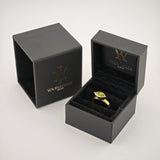 Offene schwarze Schmuckbox mit Goldring und grünem Marquise-Edelstein auf weißer Fläche.