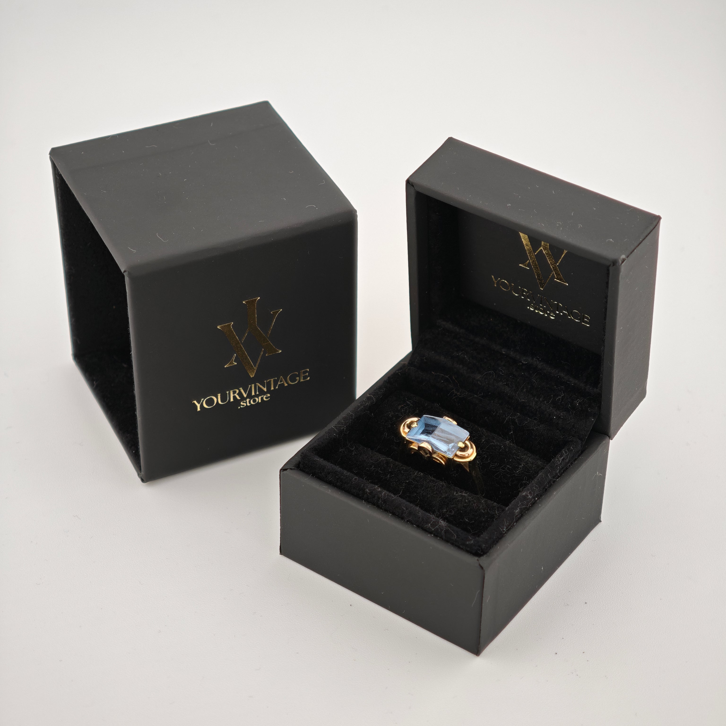 Zwei elegante schwarze Schmuckkästchen mit goldenem W-Logo, eines geöffnet mit goldenen Ring und blauem Edelstein.