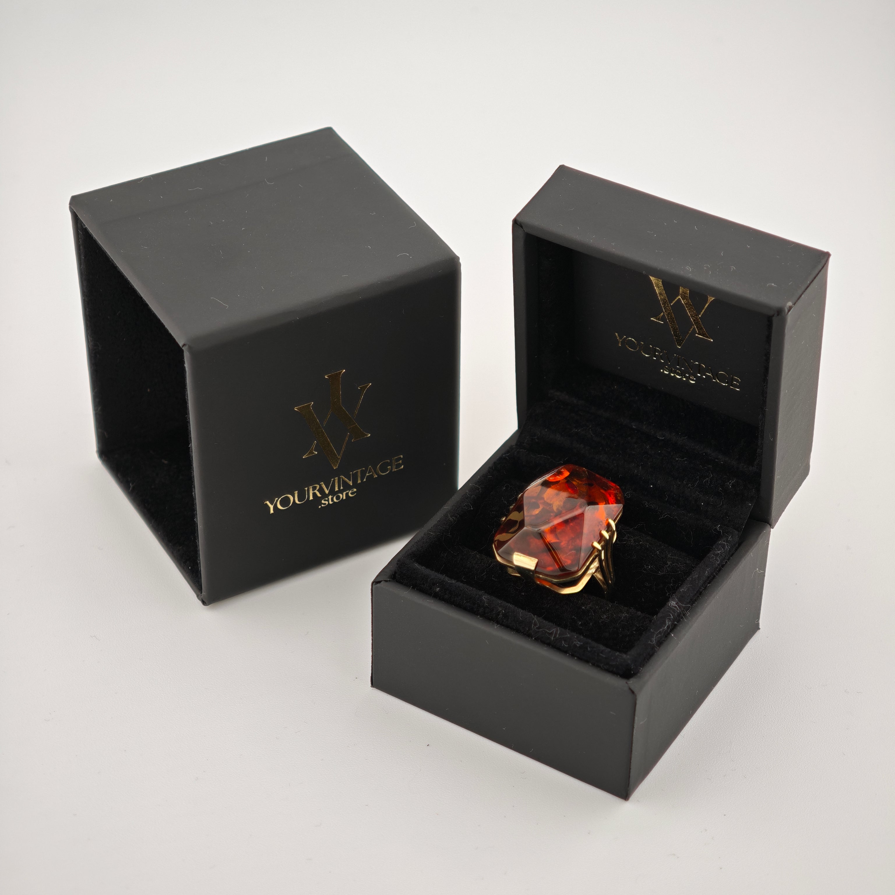 Schwarze Schmuckschachteln mit goldener Emblemprägung und Ring aus Gold und Bernstein auf schwarzem Samt