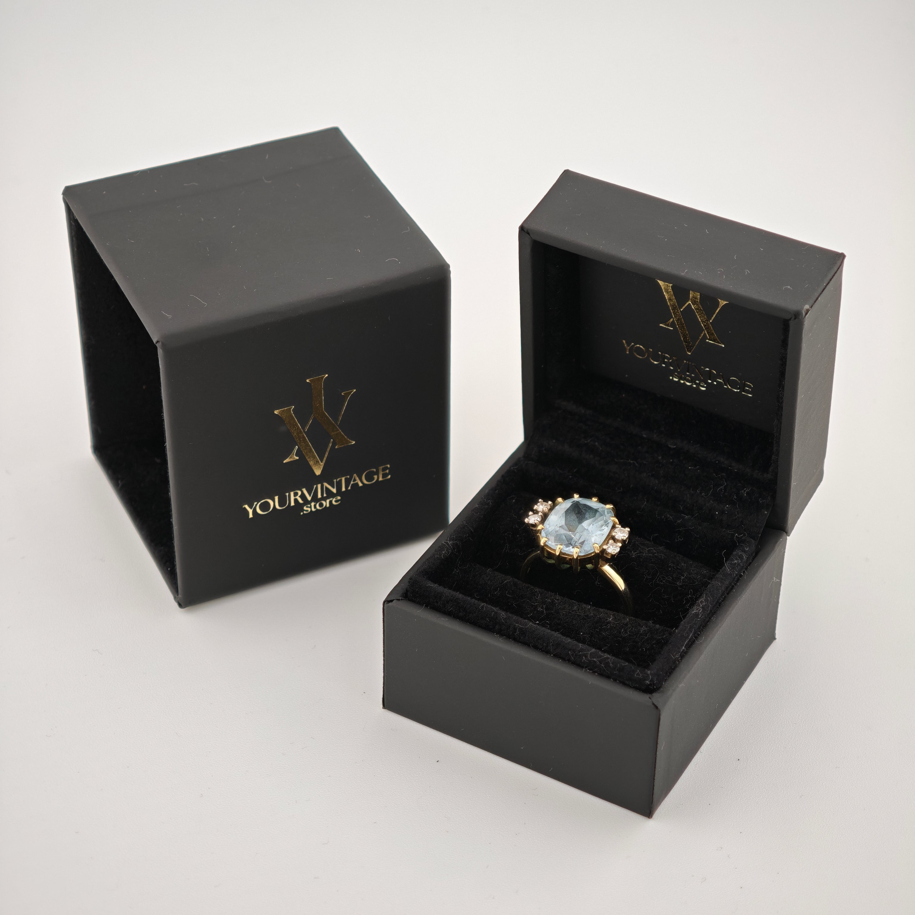 Zwei schwarze Schmuckschatullen, eine geschlossen mit goldener Aufschrift 'YourVintage Store', die andere offen mit einem Goldring und blauem Edelstein.