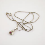 Silberne Halskette mit abstraktem Anhänger und rotem Edelstein auf weißem Hintergrund.