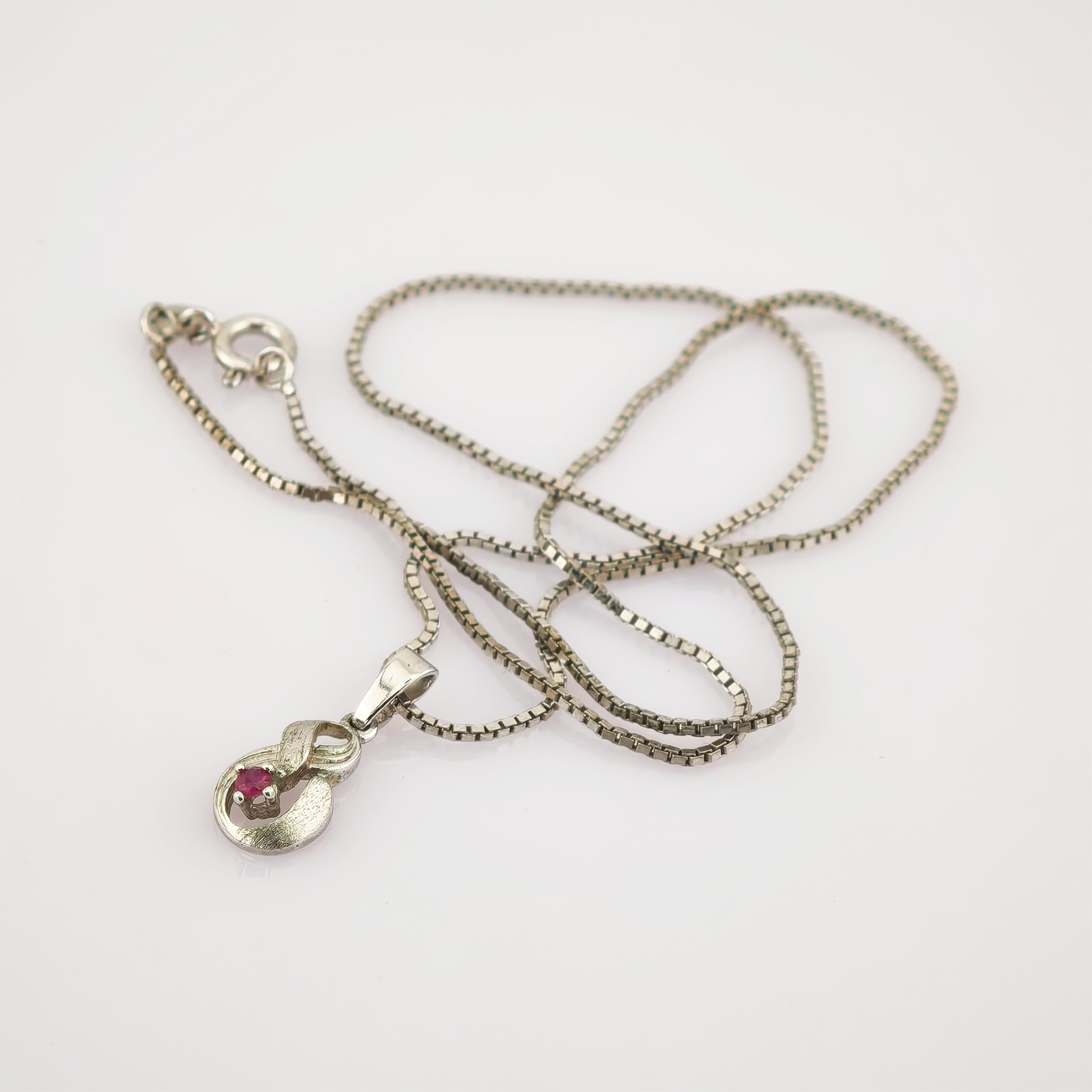 Silberne Halskette mit abstraktem Anhänger und rotem Edelstein auf weißem Hintergrund.