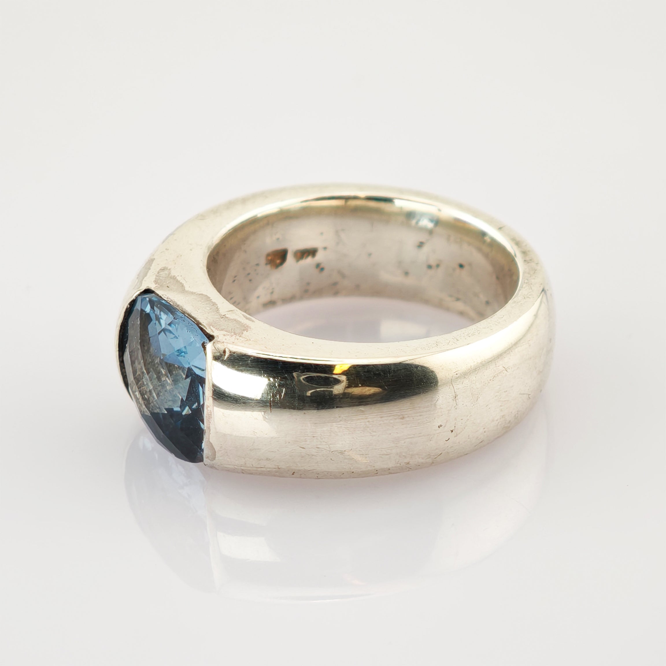 Silberner Ring mit breitem Band und großem facettiertem blauem Edelstein auf reflektierender weißer Oberfläche.