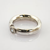 Silberner Ring mit dickem, abgerundetem Band und klarem Edelstein auf reflektierender weißer Oberfläche.