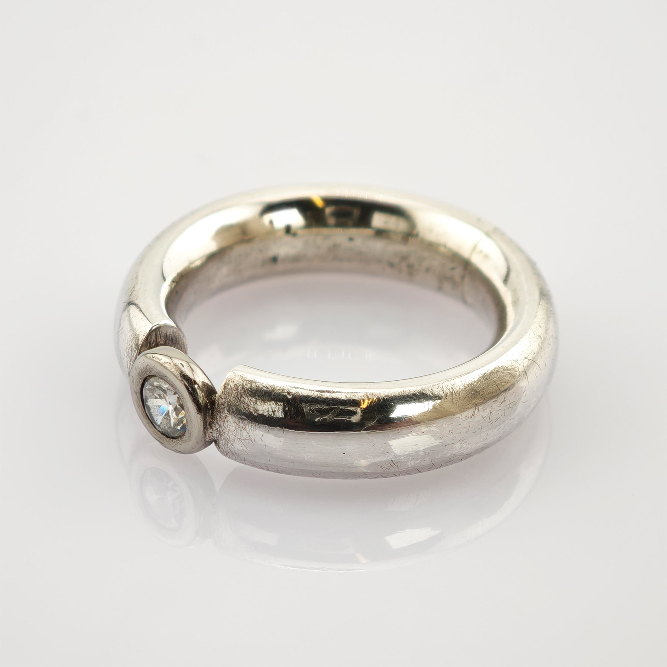 Silberner Ring mit dickem, abgerundetem Band und klarem Edelstein auf reflektierender weißer Oberfläche.