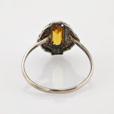 Silberner Ring mit rechteckigem gelbem Edelstein und verzierter Fassung auf reflektierender Oberfläche