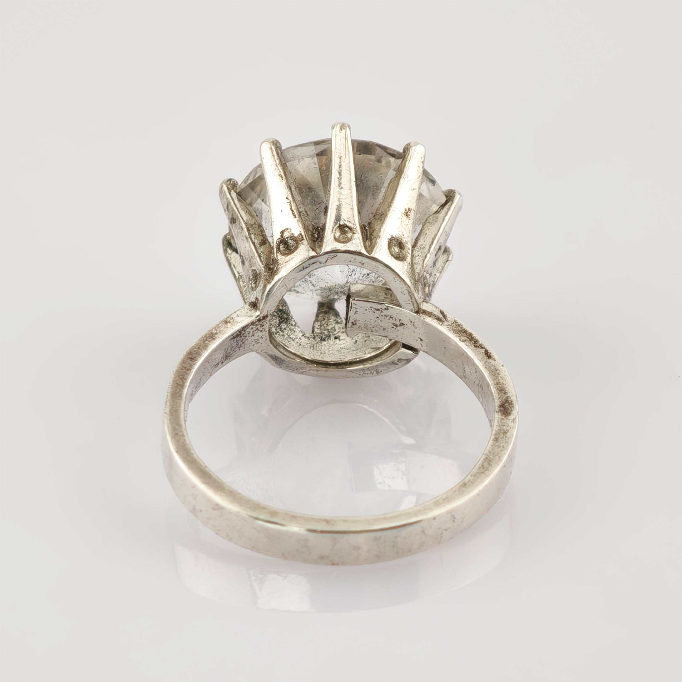 Silberner Ring mit großem klarem Edelstein in Kronenfassung auf weißem spiegelndem Hintergrund.