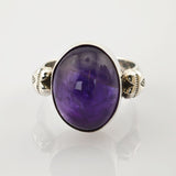 Silberner Ring mit ovalem lilafarbenem Edelstein und ornamentalem Band auf reflektierender Oberfläche.