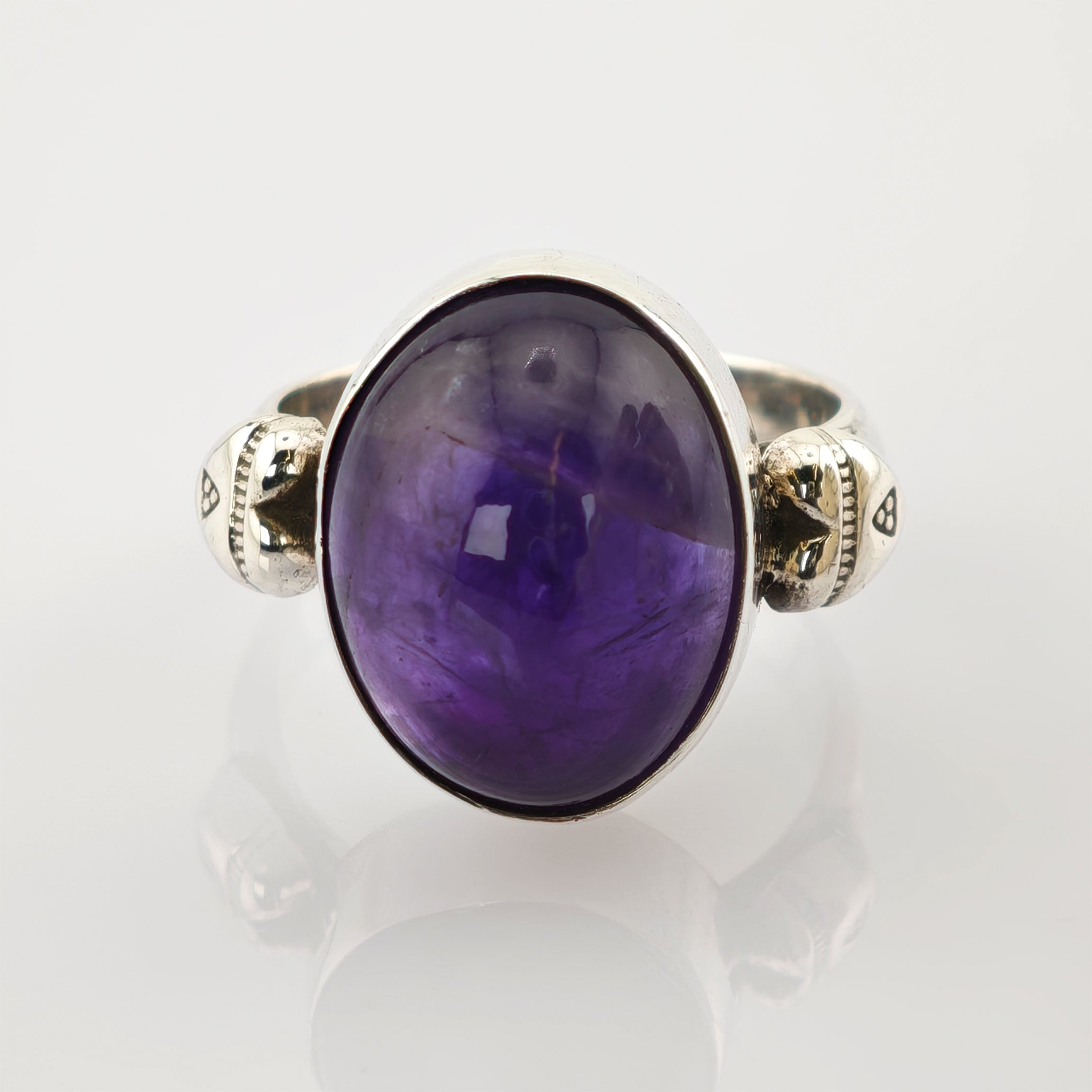Silberner Ring mit ovalem lilafarbenem Edelstein und ornamentalem Band auf reflektierender Oberfläche.