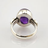 Silberner Ring mit großem ovalem violettem Edelstein und verzierten Bandseiten auf weißem Hintergrund.