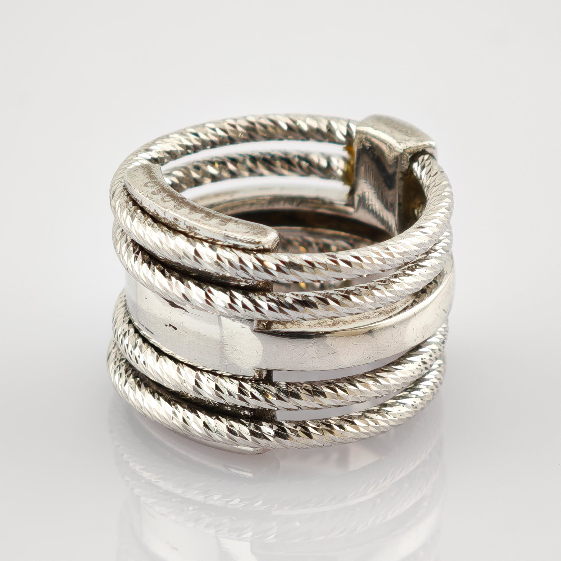Nahaufnahme eines silbernen Rings mit verflochtenen, gedrehten Bändern und glattem Mittelband auf reflektierender Oberfläche.