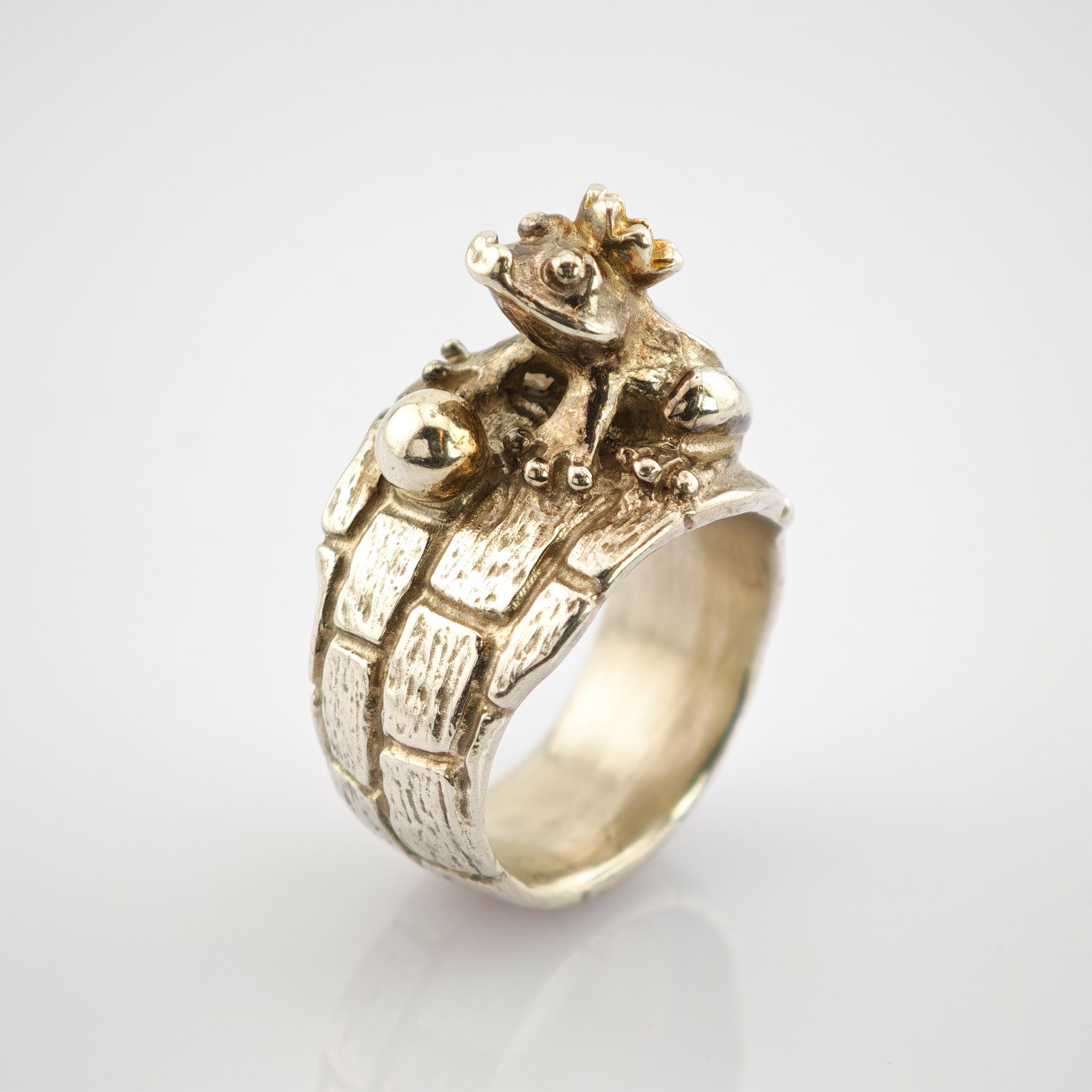 Silberner Ring mit Ziegeloptik und kleinem Frosch mit Krone neben einer glänzenden Perle.