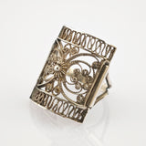 Detaillierter Silberring mit filigranem Blumenmotiv auf reflektierender weißer Oberfläche.