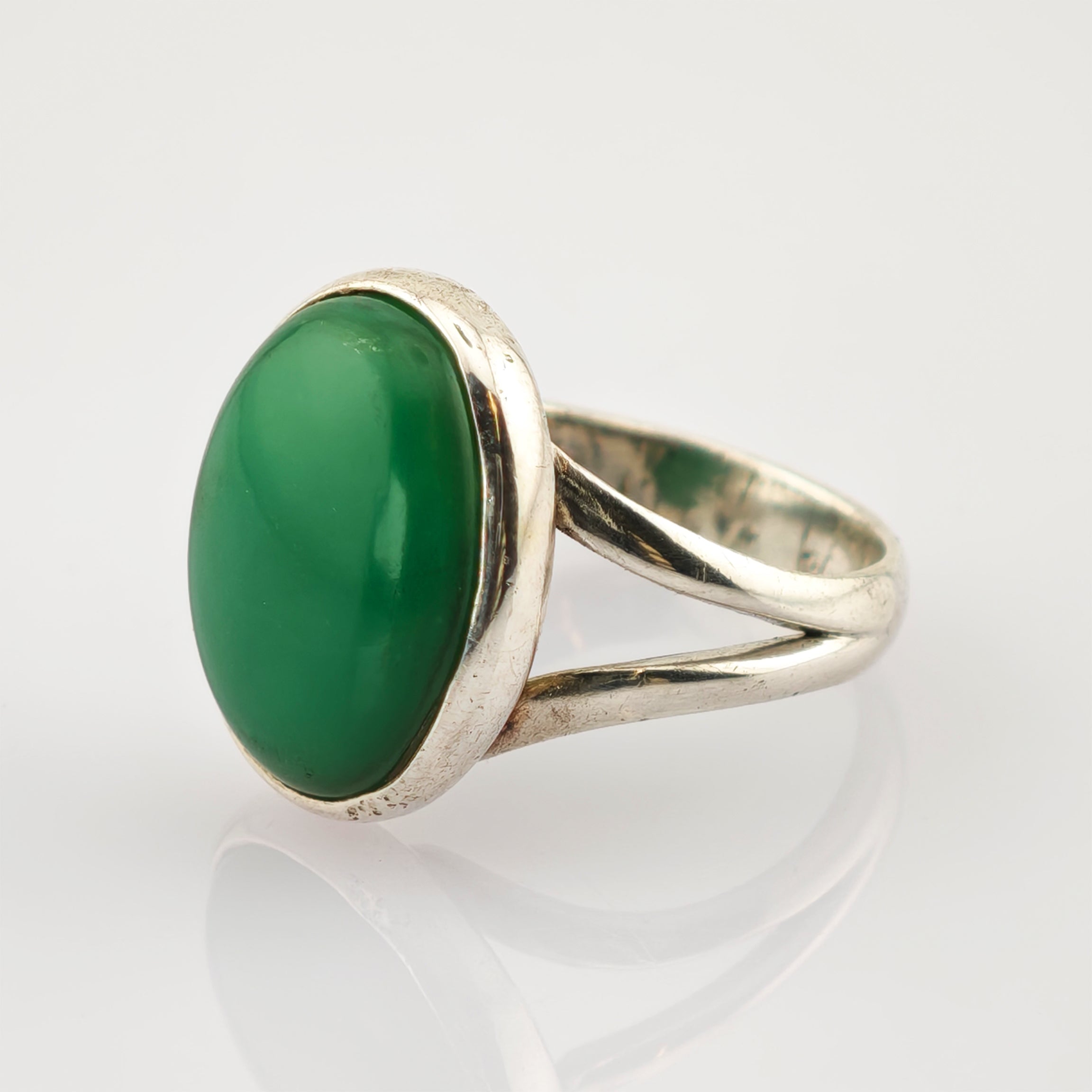 Silberring mit großem ovalem grünem Edelstein und geteiltem Ringband auf hellbeigem Hintergrund.