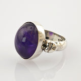Silberring mit großem ovalem violettem Edelstein und kleinem Totenkopf an der Ringschiene.