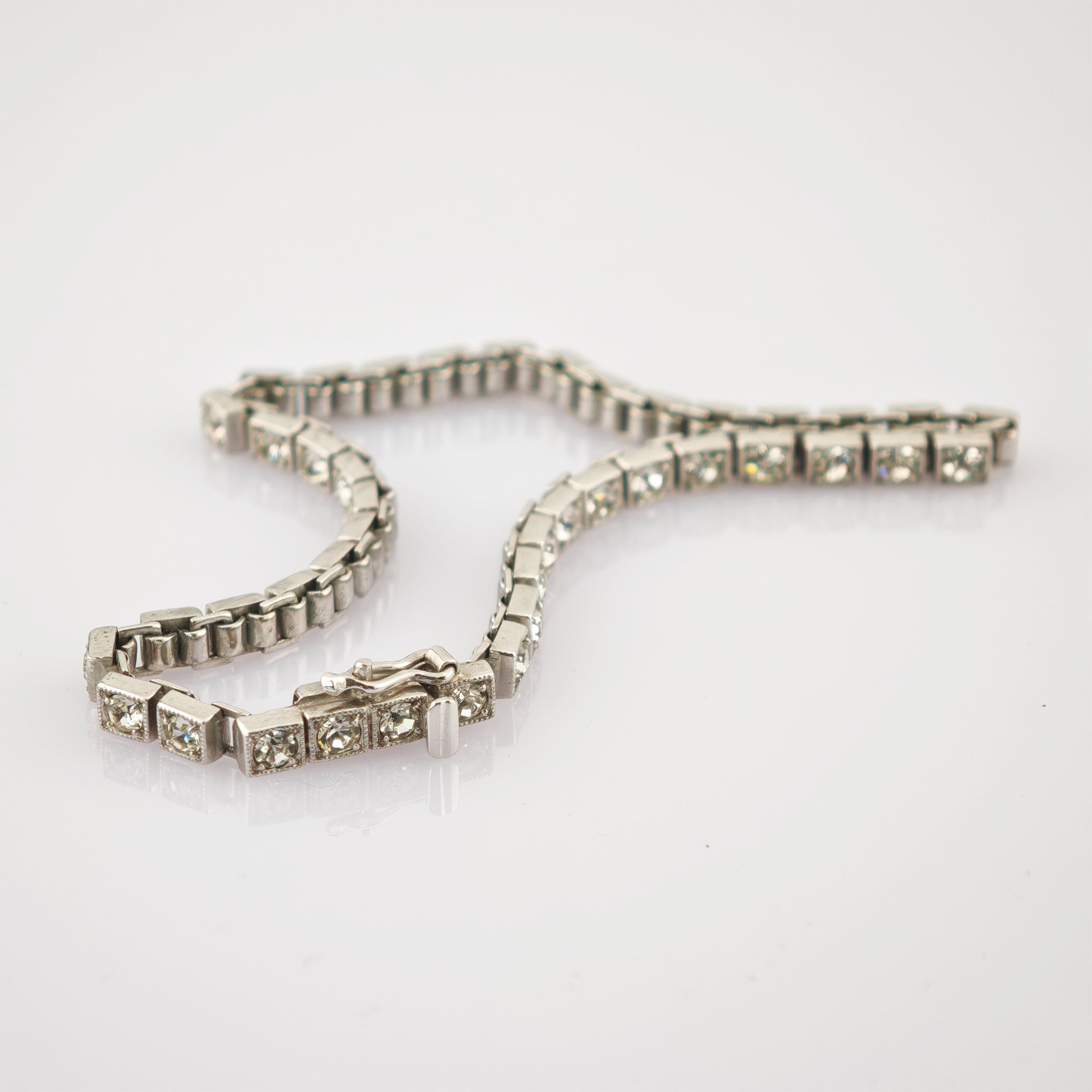 Zartes Silberarmband mit quadratischen Fassungen und funkelnden klaren Edelsteinen auf reflektierendem weißen Untergrund.