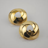 Zwei glänzende Goldohrringe mit verschlungenem Design auf reflektierender Oberfläche.