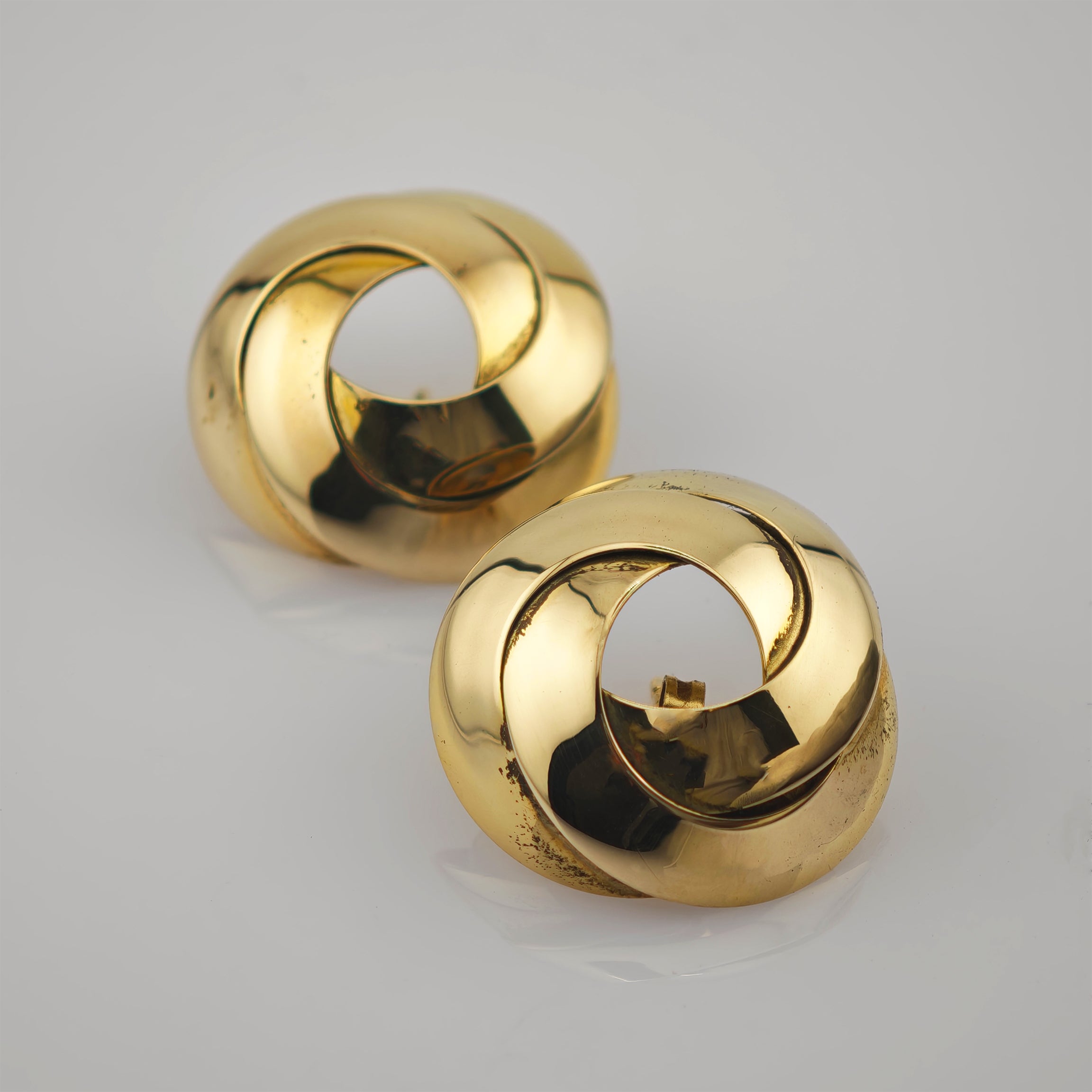 Zwei glänzende Goldohrringe mit verschlungenem Design auf reflektierender Oberfläche.